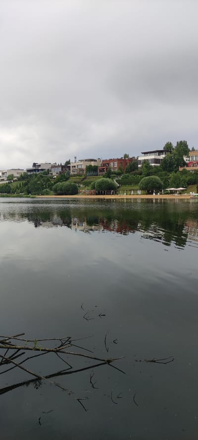 The image depicts a waterfront view in Angelovo, Russia, on a cloudy day. In the background, a row of modern houses and villas are situated on a sloping hill, interspersed with lush green trees and manicured bushes. A sandy beach with cabanas and lounge chairs is visible along the water's edge. The foreground is dominated by a dark, reflective body of water, likely a lake or river, with a few fallen branches floating on its surface. The reflection of the houses and greenery from the shore is clearly visible on the water. The overall atmosphere is serene and somewhat somber due to the overcast sky. No people are visible in the image.