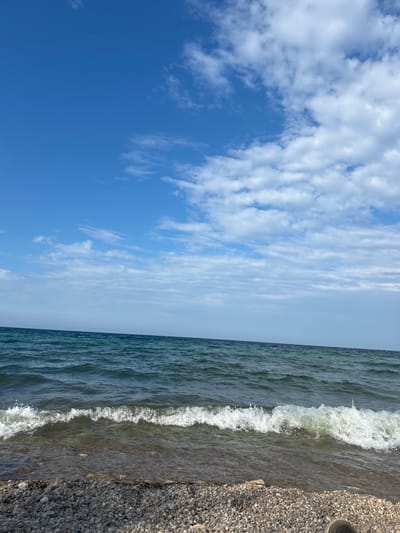 The image depicts a natural scene at the shore of a body of water, likely a lake or sea. The sky is a vibrant blue with scattered white clouds, some appearing wispy and others more substantial. The water is a deep blue-green, with gentle waves lapping against a pebble beach in the foreground. The beach is composed of small, gray and brown pebbles. The horizon line where the water meets the sky is visible. The image is a landscape view, with the water occupying the middle ground and the sky the background, and the beach in the foreground. The scene appears to be a clear, possibly sunny day. There are no people or man-made structures visible in this frame, suggesting a natural, unpopulated environment.