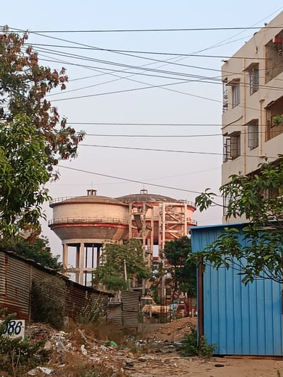This image depicts an urban environment in Anantapur, India. The central focus is on two large, elevated concrete water storage tanks. The tank on the left is a cylindrical structure with a domed roof and a red band, supported by multiple concrete pillars. The tank to its right is larger, also cylindrical, and appears to be under construction or renovation, exhibiting exposed structural elements and scaffolding.

In the foreground on the left, a section of corrugated metal fencing or shacks is visible. The ground immediately in front of these structures is heavily littered with various refuse, including plastic bags, paper, and general debris. Overgrown green vegetation is interspersed among the trash. The number "86" is stenciled in white on one of the corrugated metal surfaces.

On the right side of the foreground, a tall, bright blue corrugated metal structure, possibly a temporary cabin or storage unit, stands partially obscured by green tree foliage. Behind this structure, a cream-colored, multi-story building with multiple windows, some open, is visible.

Numerous dark utility wires, likely electrical or communication cables, crisscross the upper portion of the frame against a clear, pale sky. No individuals, actions, or specific interactions are discernibly depicted within the scene.