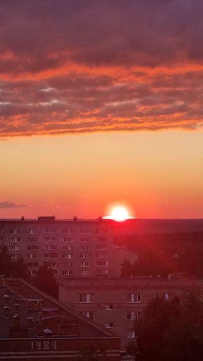 A wide-angle, vertical, outdoor, eye-level shot captures a sunset over a city. The sky is filled with colorful clouds, ranging from purple at the top to vibrant orange and red closer to the horizon. The sun, a bright white sphere with a red halo, is setting just above the city skyline. The skyline is dominated by several multi-story buildings with numerous windows. In the foreground, two lower buildings are visible, one with the numbers "1984" clearly marked on its facade. Trees are scattered throughout the urban landscape. The image appears to be taken in the city of Aleksin, Russia, given the context of prior requests.