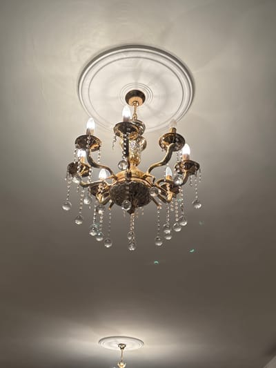 An ornate gold chandelier with crystal droplets hangs from a white, circular ceiling medallion in what appears to be a residential interior. The chandelier has six arms, each holding a bulb designed to resemble a candle flame. The ceiling itself is textured and shows some faint lines, possibly indicative of minor cracks or imperfections. Below the main chandelier, another, smaller chandelier is visible, also hanging from a ceiling medallion. The lighting suggests this is an indoor scene, likely a home or event space, and no individuals are present in the frame. The location is identified as Al Qastal, Jordan.