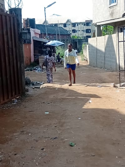 A dirt road stretches between buildings. On the left are various structures, including multi-story residential buildings and smaller shops with blue tarps. A street light rises in the background. Two figures, a woman in a patterned dress and a young person in shorts and a light-colored top, stand on the road. Debris scatters the ground. Fencing tops a gate on the lower left of the image.