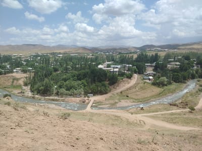 This image depicts a panoramic view of the rural settlement of Tashbulak, Uzbekistan, characterized by its arid mountainous terrain and a winding river. The settlement features a collection of low-rise residential buildings with light-colored roofs, interspersed with lush green trees and vegetation. A river flows through the valley, with a small bridge crossing it at one point. Several vehicles, including cars, a bus, and a yellow excavator, are visible in the scene, suggesting ongoing activity or infrastructure development. A few individuals are also present, walking along the riverbanks or roads. The overall landscape is dry and dusty in the foreground and on the hills, contrasting with the greener areas closer to the river and the settlement. The sky is partly cloudy, with white clouds against a bright blue backdrop.