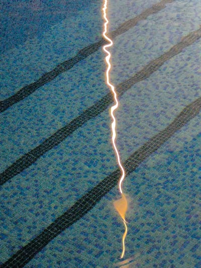 The image displays a close-up, overhead view of a blue, textured surface, likely water, in the city of Charvak, Uzbekistan. A prominent, wavy, luminous yellowish-white streak of light runs vertically through the center of the frame, broadening into a more diffuse glow at its lower end. Beneath the water's surface, four distinct, dark, diagonal stripes with a visible patterned texture, resembling tire tracks or a grid, are present. These stripes are evenly spaced and oriented from the upper left to the lower right. The background surface consists of a granular or mosaic-like pattern of dark blue and lighter blue elements. No individuals, direct actions, or interactions are visible.