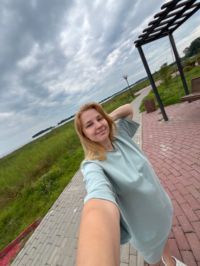 A woman with blonde hair takes a selfie outdoors in Breytovo, Russia. She is wearing a light blue t-shirt and stands on a paved walkway. To her left is a grassy area bordering a body of water. To her right, there is a wooden pergola with a swing, benches, and a trash can. The sky is cloudy.