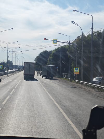 The image depicts a roadside scene in Verkhnee Sancheleevo, Russia, on a sunny day with a partially cloudy sky. The road is a multi-lane asphalt highway. In the foreground, the perspective is from inside a vehicle, as indicated by a dark object on the lower right edge, possibly a side mirror.
The primary focus is on the road and the surrounding environment. Two large trucks are visible on the road. The truck in the center is a grey semi-trailer truck facing away from the viewer. To its right, another grey box truck is parked. Several people, appearing to be two or three individuals, are standing near the parked box truck.
Traffic lights are present, with one showing a red signal. Several streetlights line the road, supported by poles and interconnected by wires. Pedestrian crossing signs with the international symbol of a person walking are also visible. One such sign is prominent on the right side of the road, accompanied by another sign with Cyrillic text. A white police car with flashing lights is parked on the right shoulder of the road, partially obscured by a metal guardrail.
The background consists of dense green trees and foliage, suggesting a rural or semi-urban setting. The road markings are clear, with white lines dividing the lanes. The overall impression is a typical daytime scene on a highway in Russia.