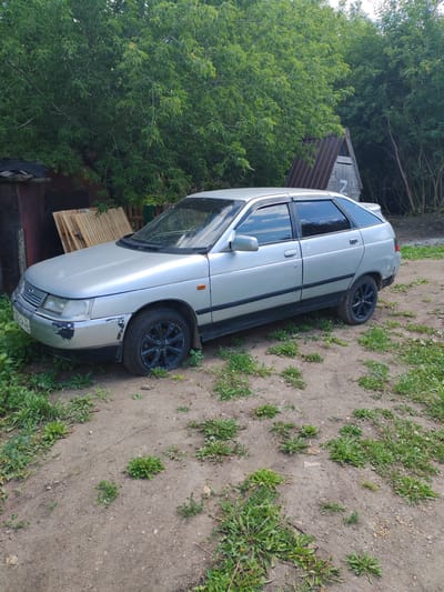 A silver hatchback car is parked on a dirt road surrounded by trees and vegetation. The car has tinted windows and black rims. The front bumper has visible damage and paint chips. To the left of the car, there is a lean-to structure with wooden planks stacked against it. In the background, a small, A-frame wooden structure with a corrugated metal roof is visible among the trees. The ground is a mixture of dirt and sparse green plants. The overall scene appears to be in a rural or semi-rural setting. The license plate of the car appears to read "K680CH 22".