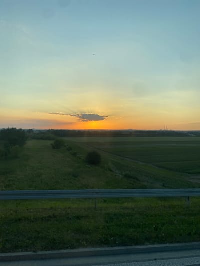 A wide shot captures a sunset over a rural landscape near Podłęże, Poland. The sky transitions from a pale blue at the top to a warm orange glow near the horizon. Rays of sunlight emanate from behind a cloud, illuminating the sky. Below the sky, a verdant field stretches across the frame, with a gentle slope. In the distance, a cityscape is faintly visible, marked by several tall structures, possibly industrial chimneys. A metal guardrail runs along the bottom of the foreground, separating a grassy embankment from what appears to be a road. The scene is serene, with no people or immediate actions depicted.