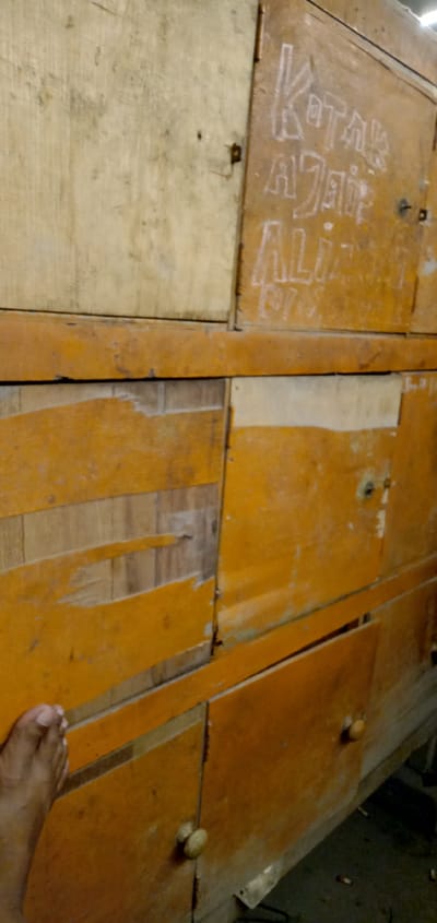An eye-level, medium shot depicts a wooden structure with a faded yellow-orange paint finish. The structure appears to be a cabinet or storage unit composed of multiple compartments with wooden doors. Some compartments are damaged, revealing the raw wood beneath the paint. The text "Kotak Ajaib Ali" is written on one of the upper compartments. A human foot is partially visible in the lower-left corner of the frame. The location is Sumput, Indonesia.