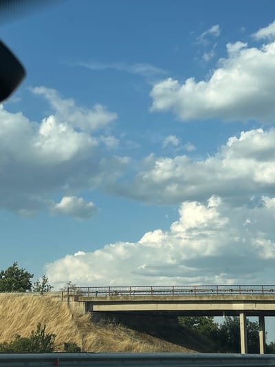 This is a vertical, slightly blurry, outdoor photograph of a bright blue sky filled with scattered white and gray clouds. On the right side of the frame, a concrete bridge structure runs horizontally, with a rusty metal railing along its edge. Below the bridge, dry, golden grass covers the landscape, with a few green trees visible near the bottom. The time of day appears to be daytime, given the bright sunlight. The weather seems clear with some clouds. The image is taken from a low angle, possibly from a vehicle, as indicated by the dark, out-of-focus object in the bottom left corner. There is no visible text in the image.
