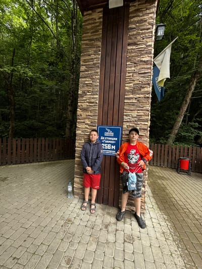 The image shows two young boys in Guamka, Russia. The boy on the left is wearing an orange jacket, a red t-shirt with a graphic design, camouflage shorts, a black baseball cap, and black Croc-style shoes. He is holding a blue and white patterned mask. The boy on the right is wearing a dark blue jacket, red shorts, and black sandals. They are both lying down or sitting on a wooden bench in front of a stone wall.

Behind the boys, a blue sign with white text reads "До станции «Гуамка» 256М" which translates to "To the station 'Guamka' 256M". Below this, it says "2 й в пути 8 минут" which means "2nd on the way 8 minutes". The setting appears to be outdoors, with trees and foliage visible in the background. The ground is paved with cobblestones, some of which are wet, suggesting it might have rained or is damp. A water bottle is visible on the ground next to the boy on the right. The overall mood seems relaxed and casual, possibly during a break from an outdoor activity. A red cylindrical object is mounted on a wooden structure in the background.