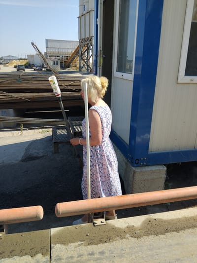 In Oliivka, Ukraine, on a sunny day, a blonde woman wearing a floral dress stands in a construction or industrial area. She is holding a long-handled implement with a white bottle attached to the end, possibly a cleaning tool or a spray bottle. The woman is positioned next to a blue and white modular building. In the background, stacks of rebar and construction equipment are visible, including a concrete batching plant. The ground is composed of dirt and gravel, and there are orange pipes in the foreground. The scene suggests an outdoor work environment under clear skies.
