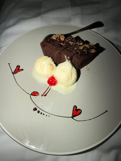 A close-up, overhead shot shows a slice of chocolate cake served on a white plate. The cake is moist and dense, topped with drizzles of chocolate sauce and sprinkled with crushed nuts. To the side of the cake are three scoops of vanilla ice cream, partially melted and pooling on the plate. A bright red maraschino cherry with a stem rests atop the ice cream.
Decorative elements adorn the plate, featuring a thin, dark brown chocolate swirl that creates abstract lines and curves. Several small, dark brown dots are placed along one of these lines, resembling a trail. At two points along the swirl, small red heart shapes are depicted, also made of chocolate.
A silver spoon is partially visible on the left side of the plate, resting on the cake. The plate is placed on a textured, white surface, possibly a tablecloth. The lighting is dim, casting shadows and highlighting the glossy finish of the chocolate and the smooth texture of the ice cream. The overall scene suggests a dessert course, likely in a restaurant or at home. The food presentation is elegant and artistic, with the chocolate swirls and heart shapes adding a touch of whimsy. There is no visible text in the image.