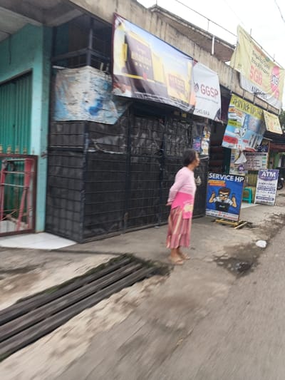 The image depicts a street scene in Cangkuang Kulon, Indonesia, featuring a commercial area with multiple shops. In the foreground, a drainage channel runs parallel to the sidewalk, covered by horizontal metal bars. To the left, a building with a teal corrugated metal rolling door and a red-framed gate is partially visible. Adjacent to it, a shop front appears closed, secured by dark, possibly black, roll-down grates or shutters.

Above the shop fronts, several large banners are displayed. From left to right, these include a partially obscured blue-and-white banner, a prominent yellow banner advertising "Sampoerna Mikaud Raya" with "Rp 18.000," a white banner with "GGSP TOKO EKA," and a yellow banner mentioning "5GB" and "LUCKY CELL." Below these, numerous smaller signs are affixed or freestanding, including a blue A-frame sign for "SERVICE HP" featuring a cartoon technician, and another sign for "ATM MINI LUCKY CELL."

An adult female individual is positioned in the mid-ground, facing generally towards the right side of the frame. She wears a light pink long-sleeved top, a pink sash with a floral pattern, and a long, striped skirt in shades of pink, white, and brown. Her footwear consists of light-colored sandals. The ground comprises concrete paving directly in front of the shops and asphalt further out in the street, both showing signs of moisture or stains. The overall impression is an active urban environment.