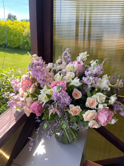 A large, lush floral arrangement is presented in a clear glass vase, its contents spilling artfully over the side. The bouquet is a symphony of pastel colors, featuring soft pink and cream roses, white alstroemeria with delicate yellow markings, and clusters of small, pale purple flowers, possibly stock or lavender, interspersed with airy sprigs of purple statice. The overall impression is one of delicate beauty and abundance.
The bouquet is positioned outdoors, on what appears to be a wooden surface, likely a table or railing, suggesting a garden setting or an outdoor patio. Behind the flowers, a translucent, ridged material, possibly polycarbonate sheeting, diffuses light and provides a softly textured backdrop. This material casts elongated shadows, hinting at the time of day. To the left and below the bouquet, a vibrant green lawn and trees are visible, bathed in bright sunlight, indicating a clear and sunny day. The presence of green foliage and the bright daylight suggest it is likely daytime, possibly late morning or early afternoon. The overall mood conveyed is peaceful and serene, enhanced by the natural light and the beauty of the flowers. There is no visible text in the image. The location context of Zaķumuiža, Latvia, aligns with the outdoor, natural setting depicted.