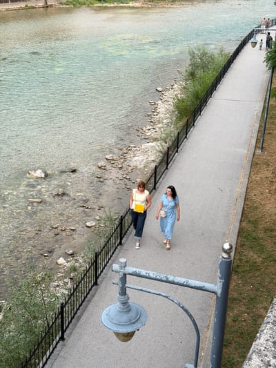 This is an overhead shot of a paved walkway alongside a shallow, clear river. The walkway is bordered by a black metal fence separating it from the riverbank, which is stony and has some greenery. On the walkway, two women are visible. The woman closer to the camera is wearing a yellow top and dark pants, holding a yellow bag, and appears to be looking down at something. The woman further up the walkway is wearing a blue dress and has a small cream-colored purse. She is facing away from the camera, towards the river. Further up the walkway, a small group of people, possibly a family, are visible standing near a lamppost. The scene appears to be a public park or riverside promenade. The lighting suggests it is daytime, and the weather seems pleasant. The location is Konjic, Bosnia and Herzegovina. No text is visible in the image.
