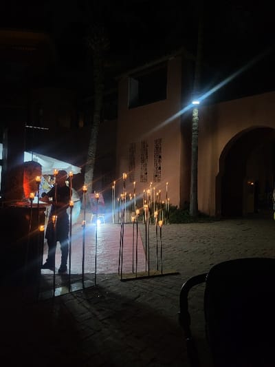 The image captures an outdoor night scene in Bouznika, Morocco, specifically within an open area or courtyard. The ground is paved with dark, rectangular pavers. In the lower right foreground, a partial, dark outline of a chair or similar furniture item is visible.

Centered in the mid-ground, a linear arrangement of approximately twenty to thirty slender, dark metal poles, each topped with a small, glowing light source resembling a candle or LED, stands upright on the pavement. These lights create a decorative illuminated path or display.

To the left of this lighting arrangement, a single individual, appearing to be an adult male, stands behind a brightly lit counter or service stall. He is dressed in dark clothing and is partially silhouetted by the light originating from the counter, with his hands positioned near its surface, suggesting an action of preparation or service. The counter includes a large, rounded, dark element on its upper left side.

The background features a structure constructed of light brown or terracotta-colored material, displaying architectural elements consistent with Moroccan design, including a prominent archway on the far right and subtle decorative patterns on its wall. Two palm trees are visible; one on the left and another on the right, directly in front of the building. A bright, wall-mounted light fixture on the right palm tree casts a strong glare and light rays across the upper mid-ground. The sky is dark and featureless.