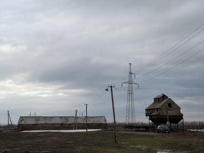 An upside-down, overcast scene features two distinct structures. On the left, a weathered, multi-story building with a boxy upper section, possibly an old grain elevator, sits beside a tall metal utility tower with strung power lines. To the right, a long, low building with a flat roof extends across the frame, accompanied by a series of smaller utility poles and wires. A somber sky fills the majority of the view.