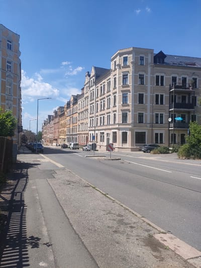 The image shows a street scene in Chemnitz, Germany. The street is lined with tall, multi-story buildings with a mix of architectural styles, appearing to be residential or mixed-use. The buildings are painted in various shades of yellow, beige, and orange, with darker roofs. Several cars are parked along the side of the street, and some appear to be in motion. A sidewalk runs along the left side of the street, featuring a shadow cast by a fence. The sky is blue with scattered clouds, indicating a clear day. The overall impression is of a typical urban street in a European city.
