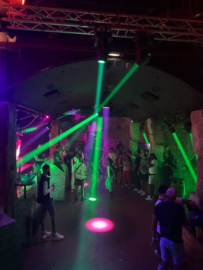 The image depicts an indoor scene, likely a nightclub or event venue, illuminated by vibrant green and purple spotlights. A group of people, primarily young adults, are gathered in the space, some standing and conversing, while others appear to be observing the surroundings. The environment features textured walls, possibly made to resemble rock or stone, and overhead lighting fixtures and trussing are visible. The floor is dark and reflects the colored lights. One individual in the foreground, wearing a black vest over a white shirt and dark pants, is facing away from the camera. Another individual in the right foreground, wearing a dark t-shirt and light shorts, is facing the camera. The scene suggests a lively, social gathering with a focus on entertainment and ambiance created by the lighting.