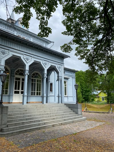 The image shows a pale blue wooden building with a covered porch supported by columns. The porch has arched openings and decorative trim. A wide set of stone stairs leads up to the entrance. To the right of the building, there is a bright yellow house visible through the trees, and a section of orange fencing. The ground in front of the stairs is paved with cobblestones and covered with fallen leaves. Trees with green foliage are prominent in the foreground and background. A cloudy sky is visible above. This building is the "Kantele" House, a museum in Sortavala, Russia, dedicated to the Karelian instrument and culture.
