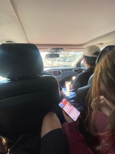 Inside a car, a driver wearing a baseball cap and black jacket is visible. Beside them, someone in a pink top and dark jacket holds a smartphone displaying a messaging app with a keyboard visible. The foreground includes a car seat, while the vehicle's interior, including the rearview mirror with hanging beads and an outside view of buildings, is also present.
