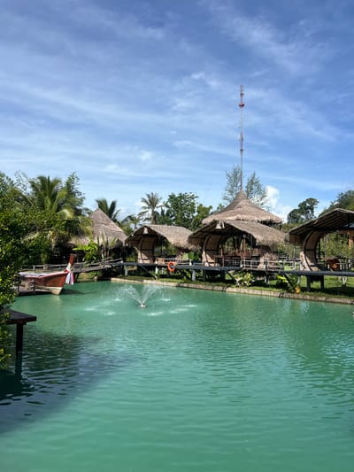 The image depicts a tranquil waterfront scene in Tambon Pa Kae Bo Hin, Thailand. A vibrant turquoise body of water dominates the foreground, disturbed by a central fountain spraying water upwards. To the left, a traditional Thai longtail boat with a red hull and wooden accents is docked, adorned with a colorful ribbon.
A wooden walkway spans the water, connecting to several thatched-roof huts or pavilions built on stilts. These structures, with their natural, rustic aesthetic, appear to be part of a resort or restaurant. Palm trees and lush green vegetation surround the huts, framing the scene with tropical foliage. In the background, a tall communication tower rises against a bright blue sky dotted with wispy white clouds. The overall atmosphere is one of serene, tropical beauty.