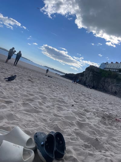 This image depicts North Beach in Tenby, Pembrokeshire, Wales, United Kingdom, under a bright blue sky with scattered white cumulus clouds. In the foreground, a pair of off-white slide sandals and a pair of dark blue Crocs clogs rest on the rippled, light-colored sand. To the left, a dark-feathered pigeon stands on the beach. Further into the mid-ground, a male figure wearing a cap, dark hoodie, and shorts holds an object while standing next to a female figure dressed in a dark hoodie and trousers. Multiple smaller figures are dispersed across the sandy expanse, indicating pedestrian activity. The right side of the mid-ground features a dark, rocky cliff face. Above this cliff, a row of multi-story white terraced buildings with numerous windows is prominently visible. The sea extends across the left background, meeting a distant headland. A faint aircraft contrail is visible high in the sky.