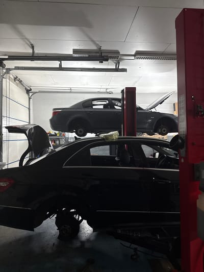 This media file shows two dark-colored cars on a car lift in a garage. The image is rotated 90 degrees counterclockwise, so the scene appears on its side. The car closer to the viewer is dark, with its hood and front bumper removed, revealing internal components. The other car, further away, is also dark and appears to be a hatchback with its rear lifted. Some of the wheels are visible, with one rear wheel of the further car clearly exposed. The garage walls are white, and there are some metal structures and piping visible along the walls and ceiling. A red car lift structure is prominent at the top of the image, supporting the cars. The lighting suggests an indoor setting, likely a workshop or service center. There is no visible text in the image. The scene appears to be an automotive service area, with the cars being worked on or prepared for repair.