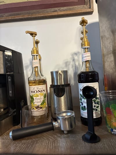 The image shows a kitchen counter with various items.  Two syrup bottles, one labeled "Blueberry" and another "French Vanilla" by Monin, have pumps attached.  A stainless steel portafilter and grinder are centrally placed. A black coffee maker is visible in the bottom portion, while a jar with fruit slices is situated at the top edge.  A framed picture is visible on the left side. The counter appears to be made of a light-colored wood.