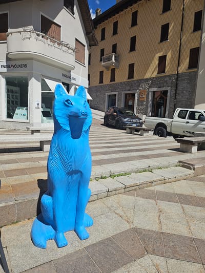 This image shows a bright blue, stylized fox statue placed on a stone-tiled plaza in Ponte di Legno, Italy. The statue is positioned in the foreground, to the left of the frame. Behind the statue, a street is visible with two buildings. The building on the left has a white facade with "ENGEL & VÖLKERS" signage and a curved balcony. The building on the right has a yellow facade with a repeating diamond pattern and stone lower walls. Several cars are parked on the street, including a dark gray SUV and a white pickup truck. The plaza is paved with large square and diamond-shaped tiles in varying shades of gray and brown. Greenery is present along the edges of the plaza and in cracks between the stones. The sky is blue with some clouds.