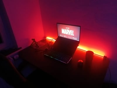 The image shows a desk setup in a dimly lit room, with red and purple lighting emanating from the walls and a light strip behind the laptop. The desk surface is dark and reflects some of the lighting.
On the desk, there is a laptop with the MARVEL logo displayed on the screen, along with the time "23:20". Below the screen, a keyboard with multi-colored RGB lighting is visible. To the right of the keyboard, there is a dark-colored cup or container and a red apple.
To the left of the laptop, there is a small tripod with a device attached. Further to the left, a portion of a chair with dark upholstery is visible. The wall behind the desk is painted in gradient colors, transitioning from pink to purple. A red light strip runs horizontally along the back of the desk, casting a glow on the wall and the desk surface.
The image captures a personal workspace, likely set up for entertainment or gaming, given the RGB keyboard and ambient lighting. There are no people visible in the image. The location is not specified to be Oberviechtach, Germany.