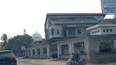 The image depicts a street scene in Meunasahlhok, Indonesia, featuring a mosque with a distinctive turquoise and white patterned dome and a minaret. The mosque is partially obscured by lush green trees. To the right of the mosque, a multi-story building, appearing to be under construction or unfinished, is visible. It has a gray concrete facade and a sloping tiled roof. Adjacent to this building is a smaller, white structure with a dark tiled roof and multiple windows. In the foreground, a man and a woman are riding a motorcycle. The man is wearing a white cap and a patterned shirt, while the woman is wearing a headscarf and a striped top. They are positioned on a paved road. To the left of the road, a black car with a partial license plate reading "BL 1447 ZL" is visible. A concrete fence with rectangular openings separates the road from the buildings and foliage. A sign with Arabic script and Indonesian text, indicating "DAYAH RAUDHATUL PUTRI AL," is mounted on a pole in the background. The sky is overcast.