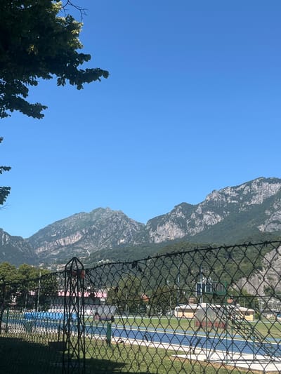 This image captures a landscape scene from Lecco, Italy, on a clear, bright day. The composition is dominated by a vast, bright blue sky on the left, providing a sense of openness and good weather. To the right, a rugged, rocky mountain range rises dramatically, its slopes covered in dense green vegetation, suggesting a natural and mountainous environment.
In the foreground, a black chain-link fence runs diagonally across the frame, partially obscuring the view of what lies beyond. Through the fence, a glimpse of a sports facility is visible. This includes blue and light blue running tracks, green grass fields, and some buildings with light-colored facades and red roofs, possibly changing rooms or administrative buildings associated with the facility. A green structure, possibly a scoreboard or information display, is also discernible.
The presence of trees with green leaves in the lower left corner and scattered throughout the background adds to the natural beauty of the scene. The lighting suggests it is daytime, likely mid-morning or afternoon given the intensity of the sunlight and the clear sky. There are no people or specific activities clearly depicted, giving the impression of a serene, perhaps early or late in the day, moment. The overall mood is one of natural grandeur juxtaposed with man-made recreational infrastructure, seen from behind a protective barrier. No discernible text is present in the image.