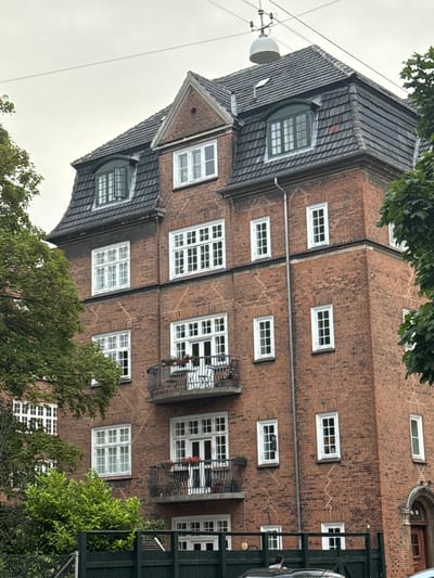 The image shows the facade of a red brick apartment building with a mansard roof covered in grey tiles. The building has numerous white-framed windows of various sizes, some with multiple panes. Two balconies with decorative wrought-iron railings are visible on the upper floors. Lush green trees partially obscure the building on the left and right sides. A dark green fence runs along the bottom of the image, with a portion of a car and two people visible behind it. The scene appears to be on a cloudy day. This building is located at **Solsortevej 16, Frederiksberg, Denmark**.