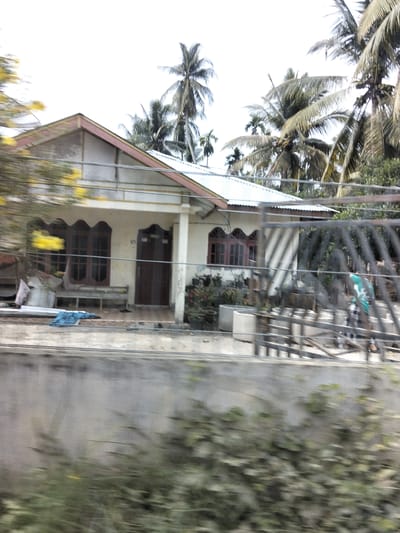 A slightly blurry, medium shot captures a single-story house in Teupinreudeup, Indonesia, under a bright, overcast sky. The house, painted a light cream color, features a white metal roof with a light pink trim. A dark brown wooden door is visible on the right side of the porch, which is supported by a white pillar. To the left of the door, there's a dark brown window with a decorative arched top and multiple panes. A wooden bench sits in front of this window. In front of the house, a grey concrete wall runs horizontally, and beyond it, a dark metal fence encloses the property. Beyond the fence, the blurred image shows a person wearing a green shirt and checkered pants. Lush green foliage and several palm trees are scattered throughout the scene, indicating a tropical environment. The overall impression is of a modest, lived-in home set against a backdrop of natural tropical elements.