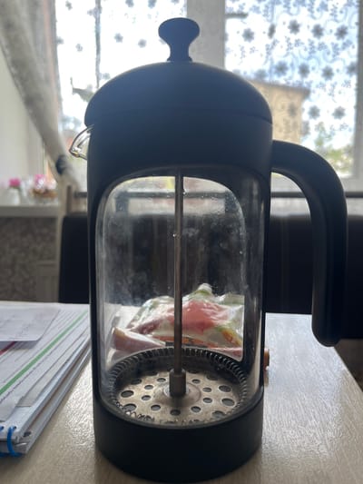 The image shows a French press coffee maker with a black handle and frame, and a glass carafe. Inside the carafe, there is a sealed plastic bag containing what appears to be fruit pieces, possibly for making infused water or a fruit tea. The French press is positioned on a light-colored, textured surface, likely a table or countertop. In the background, a window with white floral patterned curtains is visible, allowing diffused daylight to illuminate the scene. A stack of papers or a notebook lies to the right of the French press, with some handwritten text partially visible. The overall lighting suggests it is daytime. The scene appears to be indoors, in a domestic setting. No people are visible in the image. The text on the papers is in Cyrillic script and is not clearly legible.