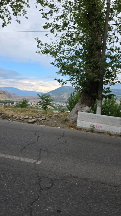 A photograph taken from a road overlooking Charvak, Uzbekistan, shows a dam and a settlement nestled between rolling hills under a partly cloudy sky. The foreground features a cracked asphalt road with a white line marking. To the right, a large tree with green leaves leans against a utility pole, and a concrete barrier with "300" spray-painted in pink is visible. The middle ground displays dry grass, scattered rocks, and a sparse arrangement of trees. Beyond this, a large dam structure dominates the landscape, with buildings and homes of the settlement clustered around it. The background is filled with layers of hills and mountains, fading into the distance under a sky with scattered clouds. The overall impression is of a slightly arid, mountainous region with significant infrastructure, possibly a reservoir or power generation facility.