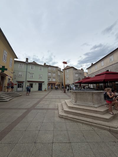 The image depicts a European town square with weathered stone buildings and cobblestone pavement. A central well, intricately carved with inscriptions, stands prominently in the foreground, surrounded by tiered stone steps. Red umbrellas offer shade over tables and chairs, suggesting an outdoor seating area for a cafe or restaurant. Several people are present in the square, some walking, others sitting and engaged in conversation. The architecture features pastel-colored facades, white-shuttered windows, and red-tiled roofs, characteristic of Mediterranean coastal towns. The sky is overcast, casting a diffused light over the scene. The overall impression is one of a historic urban space, likely in a tourist-frequented area. Specifically, this image captures the main square of Krk, Croatia, with the town's historic well serving as a central point of interest.