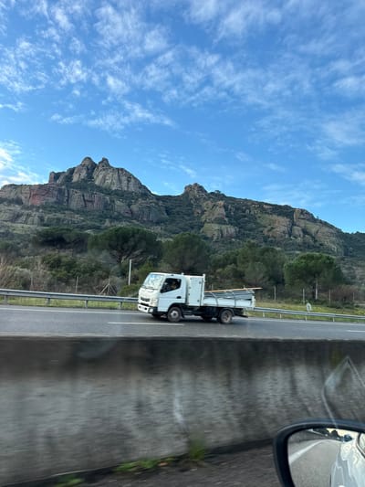 A white flatbed truck travels on a highway alongside a mountain. The mountain has a rocky face with sparse tree cover. A blue sky with scattered clouds is visible. The view is from inside a vehicle, with a side mirror in the upper right corner, reflecting the scenery. A concrete median runs along the right edge of the road.