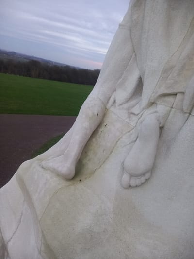 A close-up captures a white stone statue's legs and feet, draped with fabric. The statue overlooks a landscape with a clear sky transitioning into a forest and a green field, separated by a pathway. The statue's textured surface contrasts with the smooth background elements. The image appears slightly rotated.