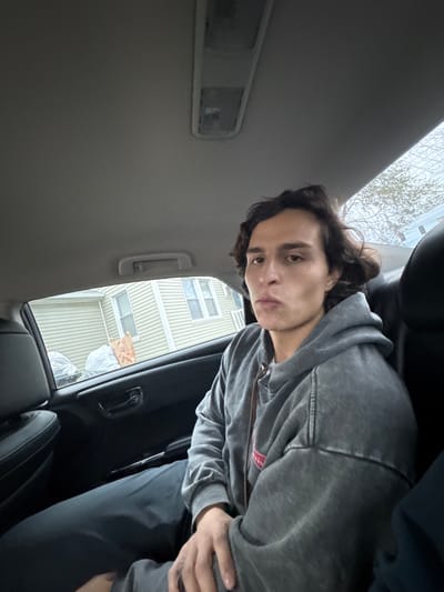 A young adult male with dark, wavy hair and a neutral expression is seated in the rear passenger side of a vehicle. He is wearing a faded grey zippered hooded sweatshirt with a small red insignia on the left chest and dark trousers. The vehicle's interior features a grey headliner, overhead dome lights, and dark door panels with a visible door handle. Through the rear passenger window, a residential backyard is visible under an overcast sky. The background includes a house with light-colored horizontal siding and two white-trimmed windows. In the yard, two large objects covered in white tarps or plastic sheeting are present, along with a brown paper bag or box on the ground. Bare tree branches are visible in the upper right quadrant of the window. The scene depicts a typical residential street setting, consistent with neighborhoods found in South Bend, United States. No specific street or address within South Bend can be identified from the image.