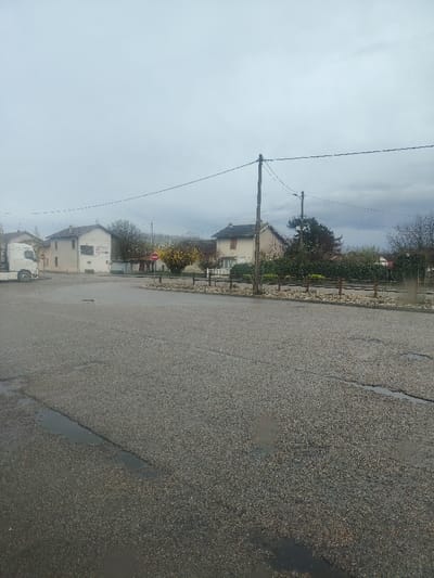 This image captures a wide-angle view of a paved area in Servas, France, under an overcast sky. The foreground features dark grey asphalt pavement with visible aggregate and multiple puddles of standing water, indicating recent precipitation. No individuals are present.

On the left side, the white cab of a semi-truck is parked, partially visible. Behind it stands a light-colored residential building with a dark roof, featuring windows and a white-painted section with indistinct markings on its facade. In the center of the scene, a small, unpaved median or island is edged with decorative light-colored rocks and low wooden posts. Within this median, a red circular no-entry sign is prominently positioned next to a yellow-flowering bush.

Further right, another light-colored residential building with a dark, sloped roof and closed shutters is visible, partially obscured by a dense line of green bushes and trees. Two wooden utility poles with overhead electrical wires extend across the midground. The sky above is uniformly grey and cloudy, without direct sunlight. The overall environment suggests a quiet, residential or semi-rural setting.