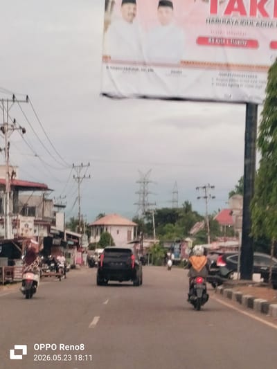 The image, taken in Teungoh Baro, Indonesia on May 23, 2026 at 18:11, shows a street scene. In the foreground, a road stretches into the distance with traffic including motorcycles and a black SUV. Pedestrians and buildings line the sides of the street, partially obscured. Overhead power lines are visible. A billboard looms above, featuring portraits of two men and text. The sky is overcast, providing ambient light.