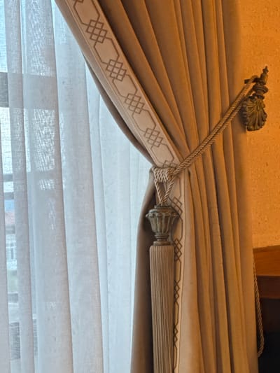A close-up shot reveals the upper portion of a window treatment, showcasing a beige curtain with a decorative trim featuring a geometric pattern. The curtain is secured by a braided rope that loops around a tassel. Behind the curtain, sheer horizontal blinds filter the light. The location in the city of Ürgüp, Turkey is not discernible from this image.