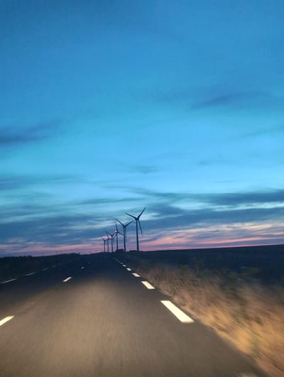 The image depicts a dusky sky with a gradient of blue and hints of pink and orange near the horizon. A line of wind turbines stands silhouetted against the sky on a slightly elevated landscape. In the foreground, a dark asphalt road stretches forward, illuminated by headlights that create streaks of light and blur the surroundings. White lines mark the edges and center of the road. The overall impression is that of driving on a highway at dusk or dawn, with renewable energy infrastructure in the distance. The scene is devoid of people or any discernible urban setting. The location is not identifiable from the image itself.