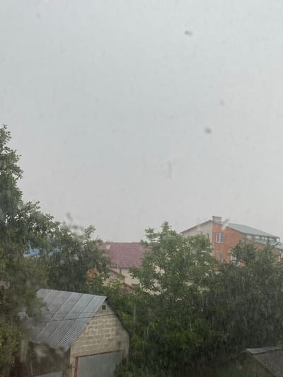 The image shows a view from a window, likely in Khust, Ukraine, on a rainy day. The foreground is dominated by smudges and streaks on the glass, indicating rain or water droplets. The background features buildings and trees. A multi-story brick building with white-framed windows is partially visible on the right side of the frame. Adjacent to it, a smaller structure with a corrugated metal roof and brick facade is also present. Lush green trees with dense foliage surround the buildings. The sky is overcast and a muted gray, suggesting either early morning, late afternoon, or a heavily clouded day. The overall mood is somewhat gloomy due to the weather conditions. There is no visible text in the image.