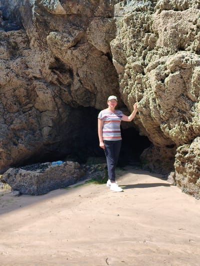 A woman stands on a sandy beach in front of a large, dark cave entrance. She is wearing a horizontally striped t-shirt, dark pants, a white baseball cap, and white sneakers. Her right hand is raised and touching the rough, textured surface of a large rock to her right. The cave mouth is a dark, shadowy opening within the rock formation. The beach is composed of light-colored sand with some scattered debris and seaweed. Sunlight casts shadows across the sand. The location is likely on the coast of Newquay, United Kingdom, given the geological features indicative of a coastal area with sea caves.