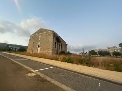 This is a wide-angle, ground-level photograph taken in the city of Shlomi, Israel, likely at dusk or dawn, given the lighting conditions and soft, diffused sky. The primary subject is a stone building with a gabled roof, possibly an old church or public building, situated on a slight incline. A prominent white cross is affixed to the upper facade of the building, indicating a religious affiliation. The building's exterior walls are constructed of rough-hewn stone blocks, and it features arched windows, some of which are boarded up or darkened. The entrance appears to be a weathered, white door.
In the foreground, a paved road with a cobblestone-like surface runs diagonally from the lower left towards the center of the frame. Adjacent to the road is a sidewalk or pathway made of rectangular, light-colored paving stones, separated by a curb. White painted lines on the dark road surface suggest parking spaces.
The area surrounding the stone building is characterized by dry, overgrown vegetation, appearing as tall, brown grasses and scattered shrubs. Behind the stone building, to the right, are modern, multi-story buildings, suggesting a transition from an older part of the city to a more developed area. The sky is a pale blue with scattered, soft clouds. The overall scene depicts a somewhat neglected historical structure juxtaposed with contemporary urban development, set against a natural landscape with hills visible in the distance. No people are visible in the frame.