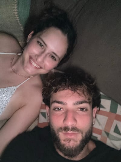 Two individuals, one female and one male, are depicted in a close-up, overhead portrait, likely an indoor selfie, in Cairu, Brazil. The female is positioned in the upper left, smiling, with dark hair tied up, wearing a light-colored sequined top with thin straps, a delicate necklace, and small hoop earrings. The male occupies the lower right, gazing directly at the camera with a neutral expression. He has dark, curly hair with lighter highlights, a full beard, and is wearing a dark t-shirt with a small earring in his left ear. They are situated close together against soft furnishings; a textured light green/beige fabric is visible behind the female, and a patterned cushion with geometric shapes in brown, beige, and light green is behind the male. The lighting is soft and even, suggesting an interior setting.